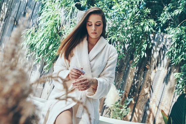 Beautiful young woman moisturizing hand cream while leaning at the bathtub outdoors