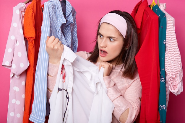 Beautiful young woman near rack with hangers. Shocked lady finds awful stain on white blouse. Brunette female holds shirt. Girl goes shopping. Girl wears sweater in mall. colorful clothes in store.