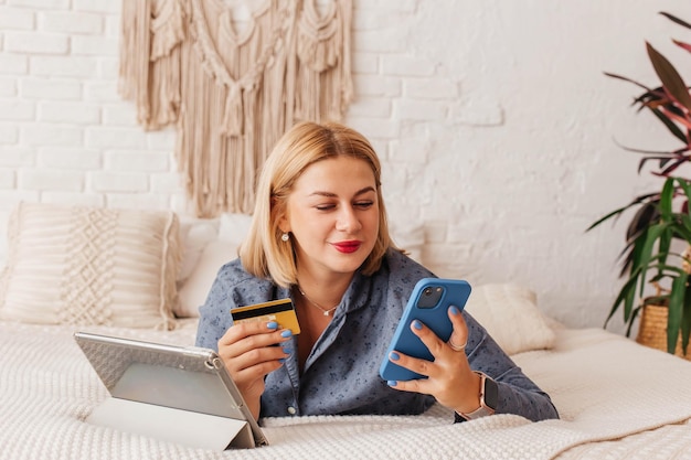 A beautiful young woman in pajamas sits on the bed with a computer and a phone makes an online payment Online shopping