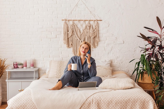 Beautiful young woman in pajamas, working on a laptop in bed, talking on the phone. Online shopping.