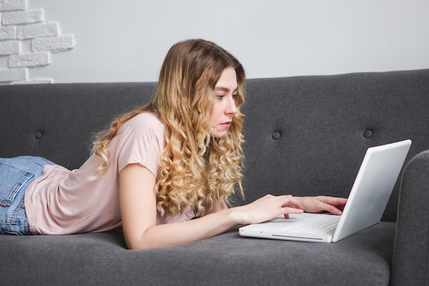 Beautiful young woman in pink shirt working on the laptop Girl lying on the sofa with notebook Dis