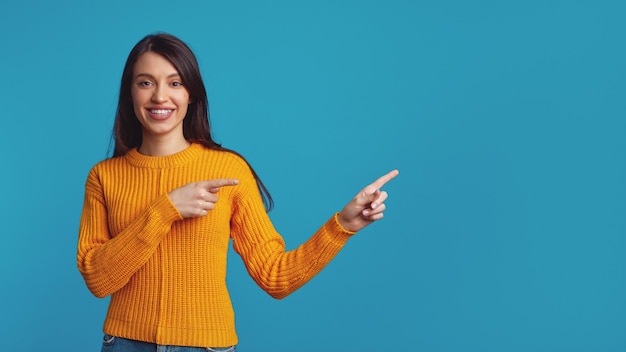 Beautiful young woman points aside on empty blue space with both