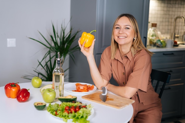 Beautiful young woman poses with yellow bell pepper at a table in kitchen with modern interior, looking at the camera. Concept of healthy eating.