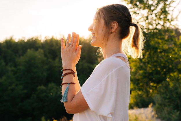 Beautiful young woman putting hands in namaste mudra posture outside in park evening on background of soft sunlight Lady with closed eyes holding hands in namaste pose