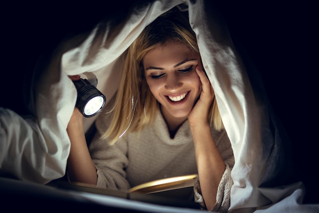 Beautiful young woman reading book under bed cover and holding a flashlight.