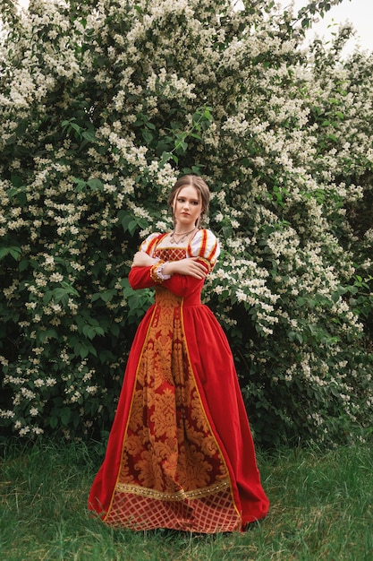 beautiful young woman in a red medieval dress is standing in the garden in white flowers