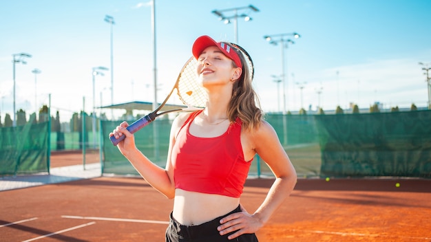 Beautiful young woman in red sports clothes posing with tennis racket at the court