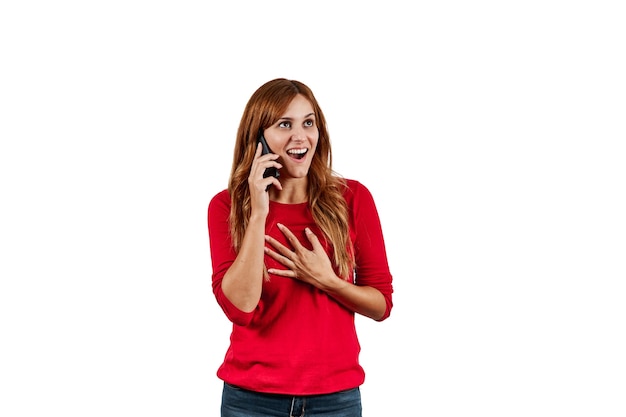 Beautiful young woman in a red sweater, surprised while talking on the phone, isolated on a white wall.