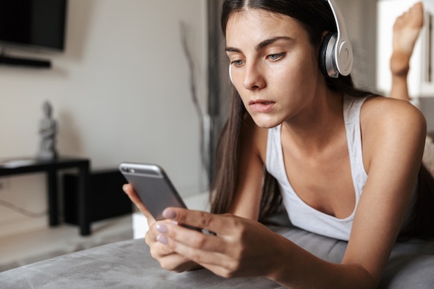 Beautiful young woman relaxing on a couch at home, listening to music with headphones, holding mobile phone