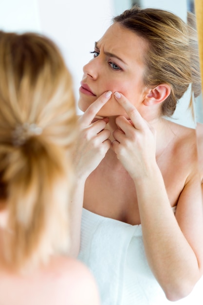 Beautiful young woman removing pimple from her face in a bathroom home. 