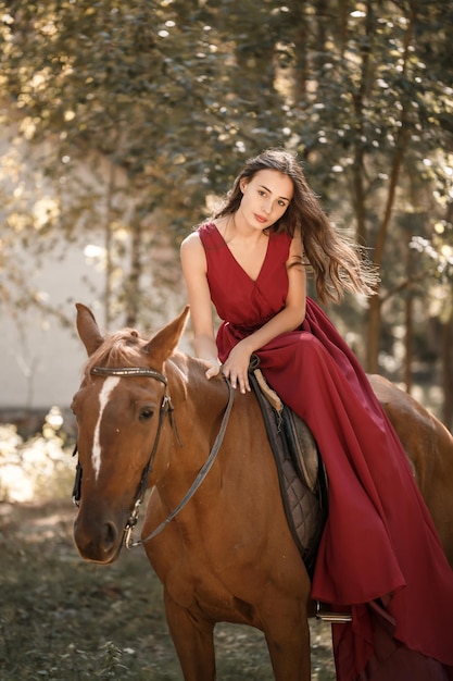 Photo a beautiful young woman in a silk dress sits astride a horse. horse ride on a sunny day in the forest.