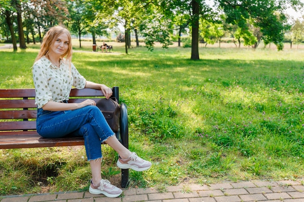 The beautiful young woman sits on a bench. Stylish woman stay in the park.