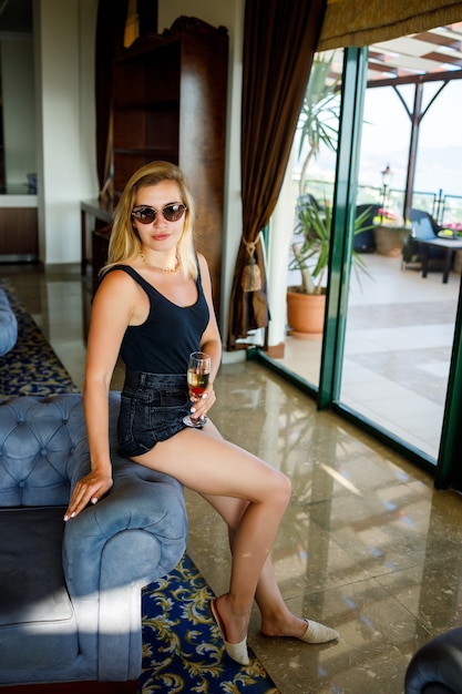 Beautiful young woman sits on a soft armchair in the hotel lobby with a glass of champagne on vacation. She is wearing sunglasses and a smile on her face. Vacation concept