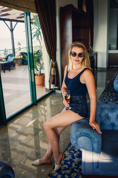 Beautiful young woman sits on a soft armchair in the hotel lobby with a glass of champagne on vacation She is wearing sunglasses and a smile on her face Vacation concept