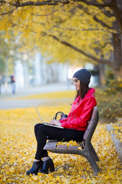 Beautiful young woman sitting in a bench and using her laptop in autumn.