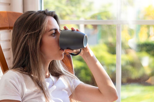 Beautiful young woman sitting drinking from a mug while thinking and looking through a window