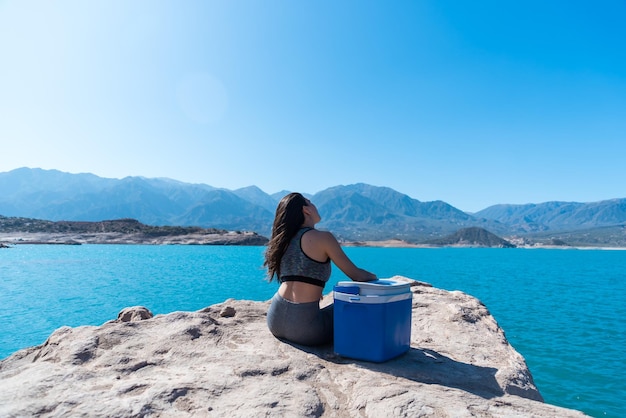 Beautiful young woman sitting on her back with a cooler on a lake between mountains