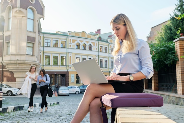 Beautiful young woman sitting outdoors using laptop city street background