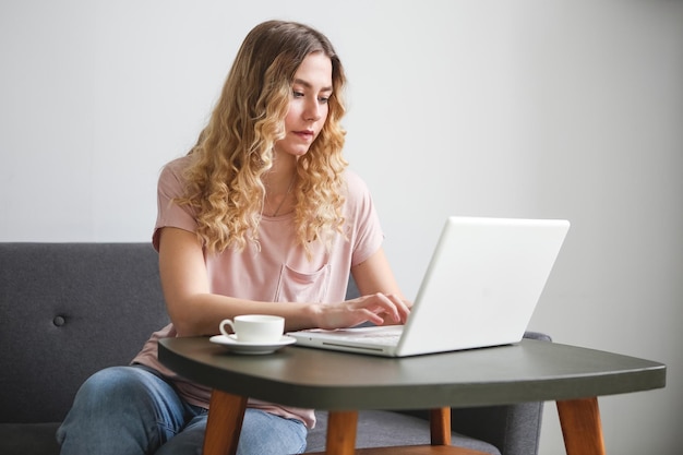 Beautiful young woman sitting on the sofa with white cup of coffee working on white laptop Girl in