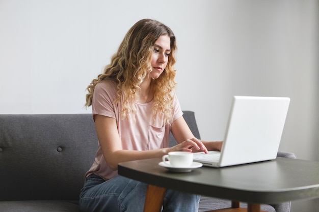 Beautiful young woman sitting on the sofa with white cup of coffee working on white laptop Girl in