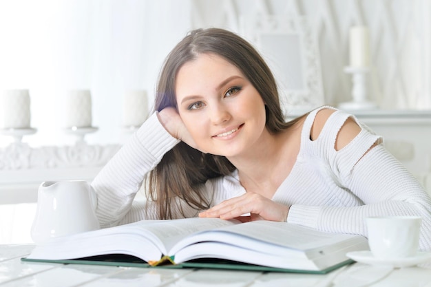 Beautiful young woman sitting at table with open book and cup of tea