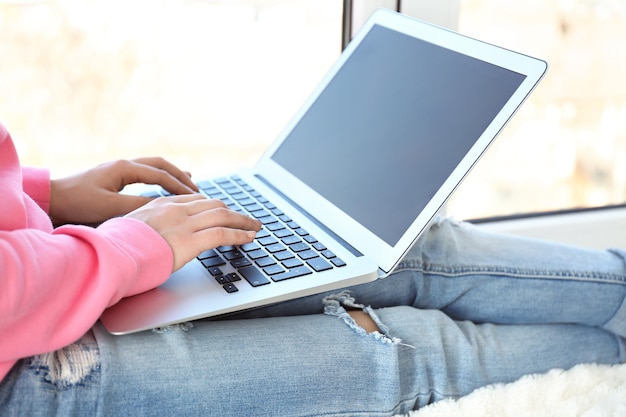 Beautiful young woman sitting on windowsill and using laptop