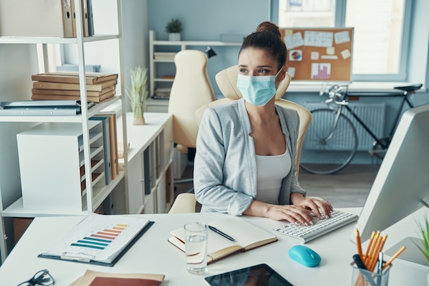 Beautiful young woman in smart casual wear and protective mask using computer