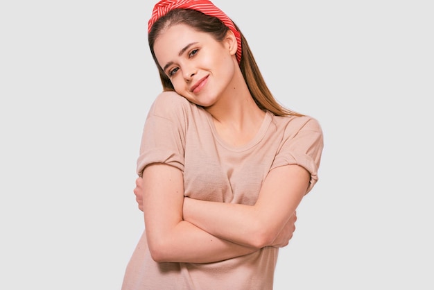 Beautiful young woman smiling feels happy keeps hands crossed posing against white background Pretty female has joyful expression wearing red head band and beige tshirt