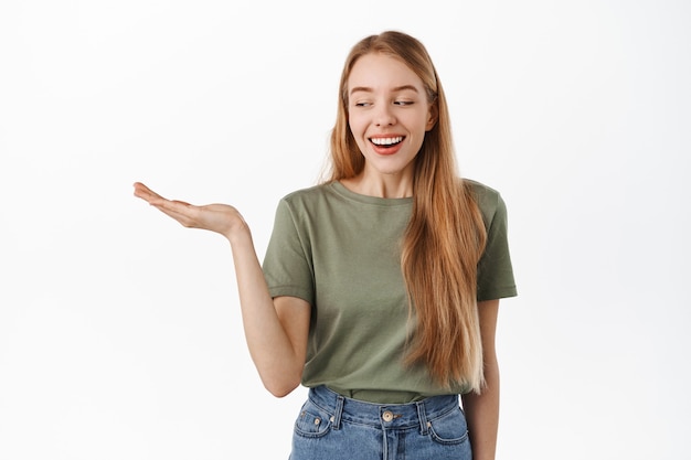 Beautiful young woman smiling, looking at her empty open hand pleased, carry an item, display advertisement product in palm, standing in t-shirt against white wall