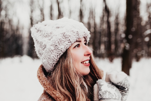 Beautiful young woman in snowy fancy winter woodland. Girl wearing fluffy gloves, cap and coat. Christmas forest, trees on blurred background. Crossed hands because of cold weather. High quality photo