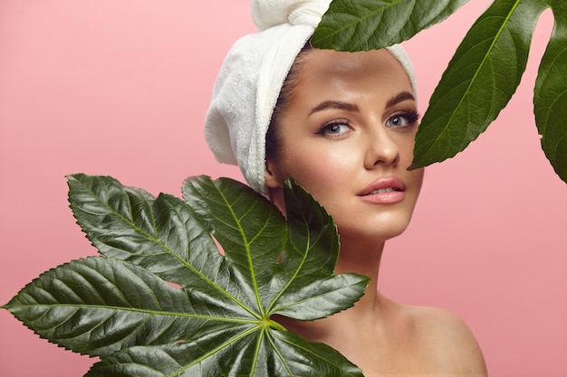 Beautiful young woman in spa turban on head stands among tropical leaves over pink isolated