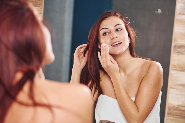 Beautiful young woman standing in bathroom, looking into the mirror and taking care of her face.