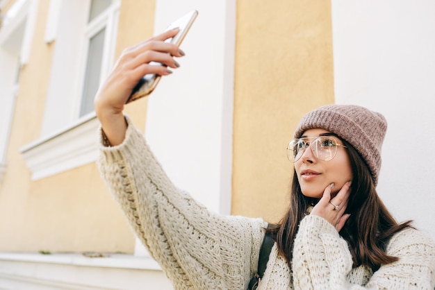 Beautiful young woman standing outdoors against building's wall and taking self portrait on smart phone Brunette female dressed in knitted sweater hat eyeglasses making selfie on the city street