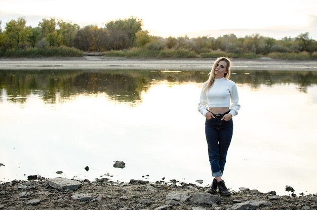 Beautiful young woman stands alone on the bank of the river in a white sweater fulllength