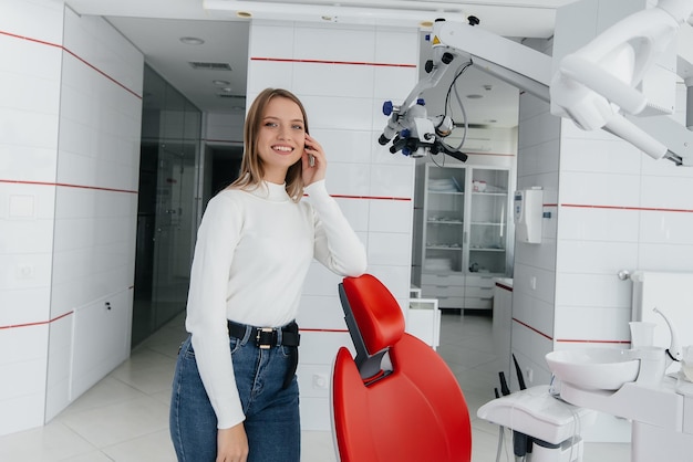 A beautiful young woman stands near a red dental chair in modern white dentistry and smiles Dental prosthetics treatment and teeth whitening