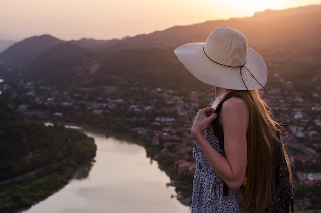 Beautiful young woman in straw hat with river and mountains on background. Sunset soft light.