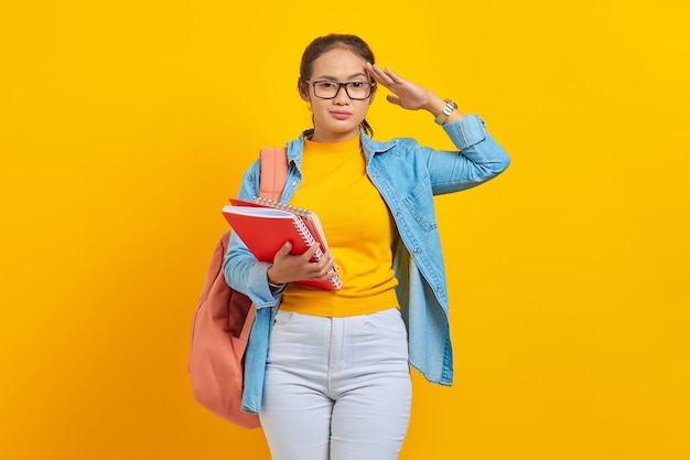 Beautiful young woman student in denim clothes with backpack holding notebook and showing respect gesture with hand isolated on yellow background Education in high school university college concept