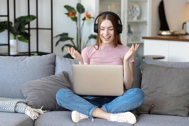 Beautiful young woman student in headphones and using a laptop for online communication sitting on the couch at home