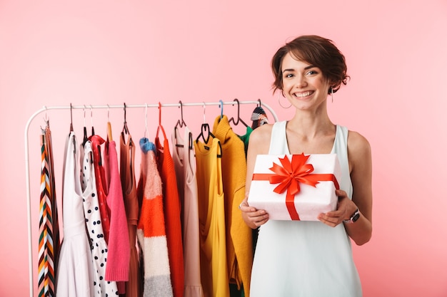 Beautiful young woman stylist standing at the clothes rack, holding present box