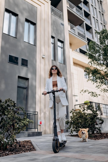 Beautiful young woman in sunglasses and white suit is riding on her electric scooter with Corgi dog near modern building in city and looking away