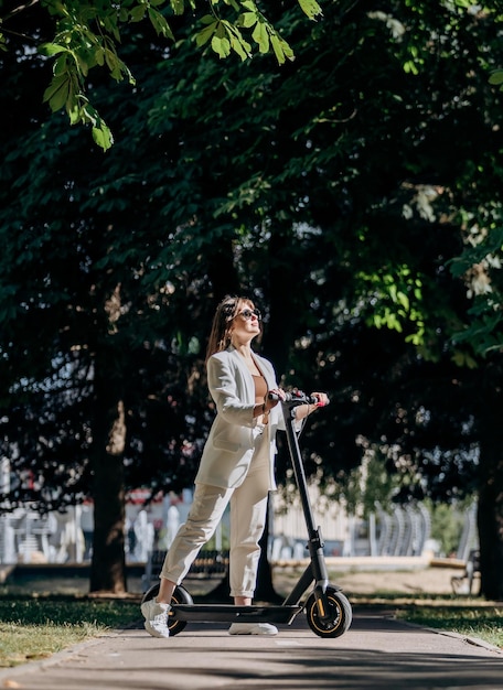 Photo beautiful young woman in sunglasses and white suit is standing with her electric scooter in city parkland