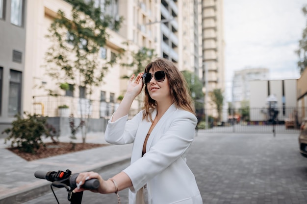 Beautiful young woman in sunglasses and white suit standing on her electric scooter near modern building and looking away