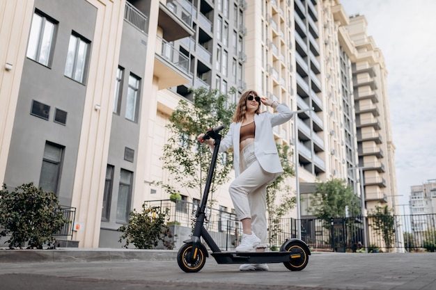 Beautiful young woman in sunglasses and white suit standing on her electric scooter near modern building and looking away