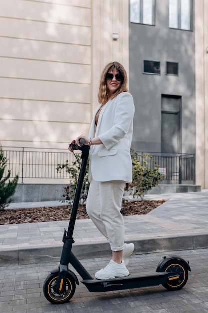 Beautiful young woman in sunglasses and white suit standing with her electric scooter near modern building in city and looking away