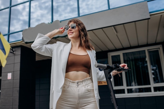 Beautiful young woman in sunglasses and white suit standing with her electric scooter near modern building and looking away