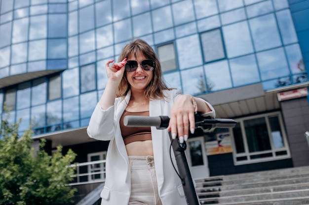 Beautiful young woman in sunglasses and white suit standing with her electric scooter near modern building and looking at camera