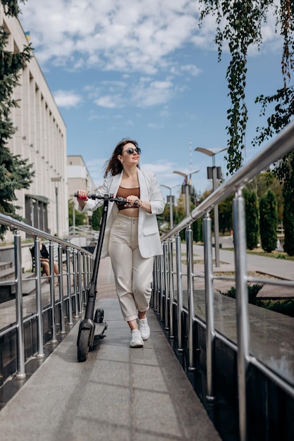 Beautiful young woman in sunglasses and white suit walking with her electric scooter near modern building and looking away