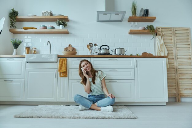 Beautiful young woman talking on mobile phone while sitting on the floor in the kitchen