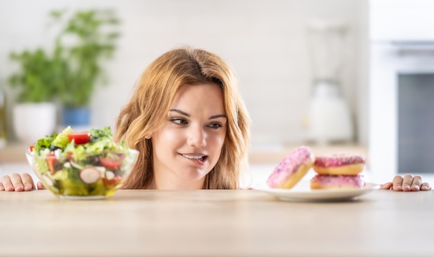 Beautiful young woman tempted having to make choice between fresh lettuce salad and doughnut