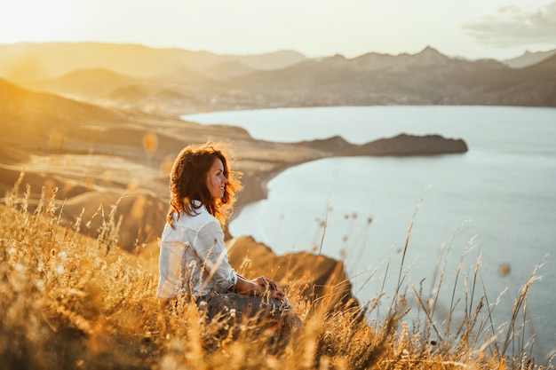 A beautiful young woman tourist is sitting on top of a mountain and enjoying the view of the sunset